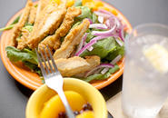 Plate of grilled chicken salad, ice water with lemon wedge, and a small bowl of fruit with a fork resting on it.