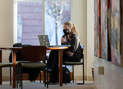 A student sits by a window on campus, working on their laptop.