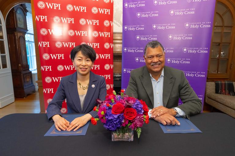 WPI President Grace Wang and Holy Cross president Vincent Rougeau sit together in front of backdrops for each institution