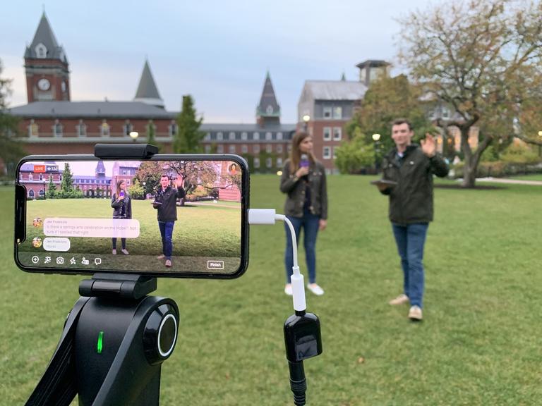 two students talking into a microphone on an open field and a smartphone recording them on a gimbal