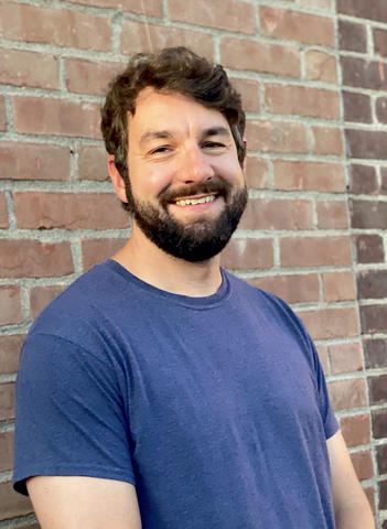 Hugh Martin, wearing a blue t-shirt, smiles and stands in front of a brick wall