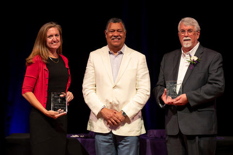 a woman in a black dress and red sweater and a man in a dark suit hold awards with a man in a light colored suit between them